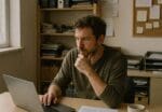 Small business owner researching at a laptop in a cluttered office back room with notes and coffee nearby