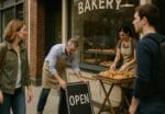 Bakery owner and employee set out an OPEN sign as customers walk past a neighborhood storefront