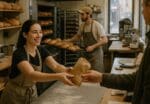 Busy local bakery staff serving a customer while bread bakes in the oven